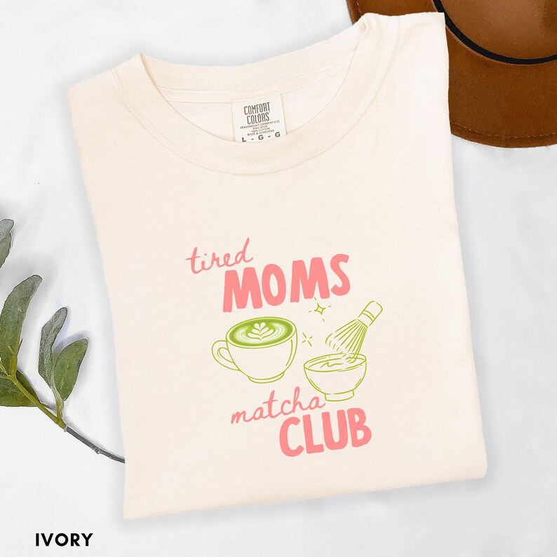 May include: Ivory t-shirt featuring the text "tired MOMS matcha CLUB" in pink and green, with a matcha latte and whisk design. The shirt is made of a soft material and is a Comfort Colors brand.