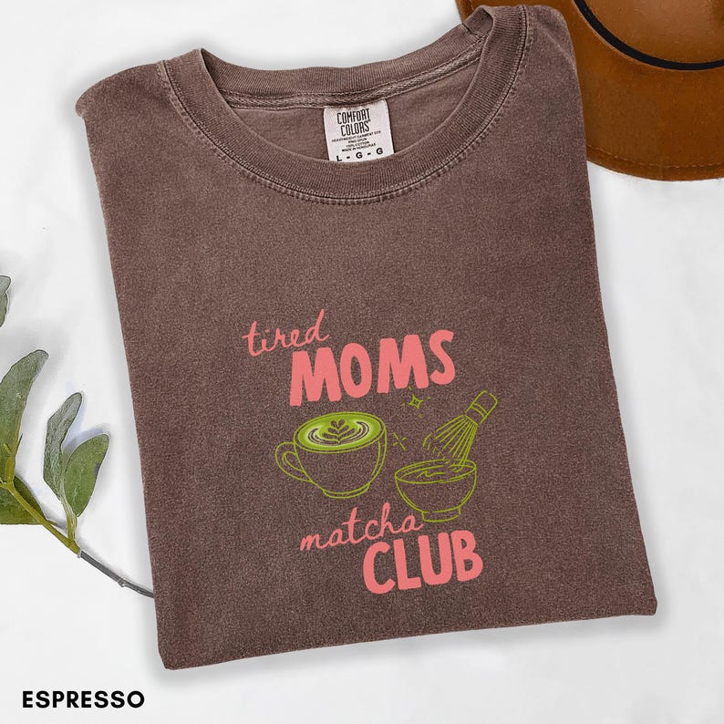 May include: Espresso-coloured t-shirt featuring the text "tired MOMS matcha CLUB" in pink and green. The design includes a matcha latte and a whisk. The shirt is a Comfort Colors brand.