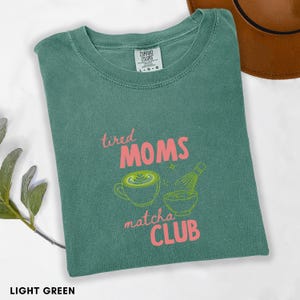 May include: Light green t-shirt with the text "tired MOMS matcha CLUB" in pink. The shirt features a matcha latte and whisk graphic in green. The shirt is folded and has a Comfort Colors tag.