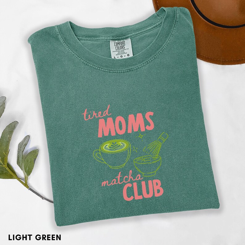 May include: Light green t-shirt with the text "tired MOMS matcha CLUB" in pink. The shirt features a matcha latte and whisk graphic in green. The shirt is folded and has a Comfort Colors tag.