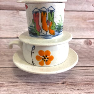 May include: A ceramic Vietnamese coffee filter set. The top filter features a painted scene of figures in conical hats and colourful robes. The bottom cup has a handle and a painted orange flower design. The set is white with speckled detailing.