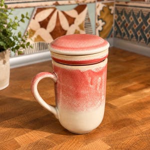 以下が含まれることがあります： 蓋付きの陶器製ティーマグ。上部と蓋は質感のあるコーラルレッドの釉薬で仕上げられており、滑らかなクリームホワイトのベースに向かって垂れ落ちるようなデザインです。側面には湾曲したハンドルが付いています。
