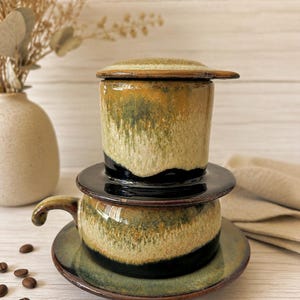 Puede incluir: Un juego de filtro de café vietnamita de cerámica que incluye una taza, un platillo, una cámara de filtro y una tapa. La cerámica presenta un esmalte reactivo con tonos mezclados de verde musgo, amarillo mostaza y negro profundo.