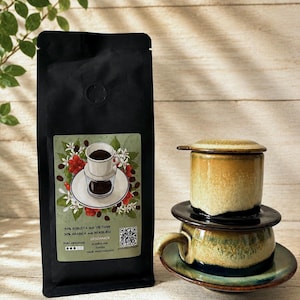 May include: A black coffee bag with a label featuring a coffee cup illustration and text, alongside a ceramic Vietnamese coffee filter set. The filter has a yellow and green reactive glaze, consisting of a cup, a lid, a press, and a saucer.