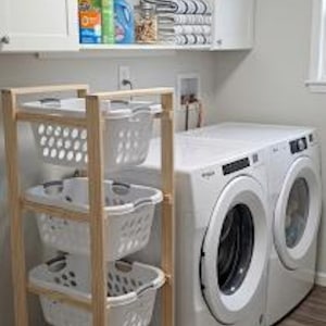 May include: A laundry room with a wooden frame laundry sorter with four white plastic baskets. A white washer and dryer are to the right. A small woven basket sits on the floor. Cabinets and a shelf hold laundry supplies.