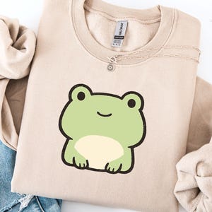 Puede incluir: Sudadera beige con un diseño de rana de dibujos animados en verde claro y crema. La rana tiene ojos negros y una boca sonriente. Un collar plateado con un colgante de sol está sobre la sudadera. La etiqueta dice "GILDAN".