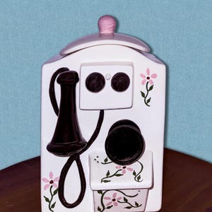 May include: A white ceramic telephone-shaped cookie jar with a lid and pink accents. The jar features a black telephone receiver, dial, and floral details. The jar is on a brown surface with a blue background.