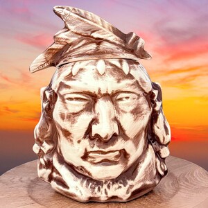 May include: A ceramic mug shaped like a human head wearing a feathered headdress. The mug features a glossy finish with a two-tone brown and cream color scheme, highlighting the facial features and the textured details of the feathers on top.