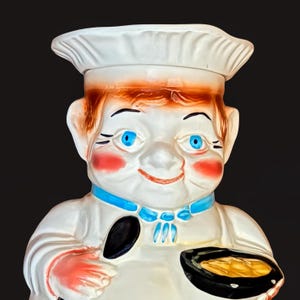 May include: A ceramic cookie jar shaped like a chef wearing a white hat and uniform with a blue neckerchief. The figure has bright blue eyes, rosy cheeks, and reddish-brown hair. The chef holds a black skillet containing a yellow food item.