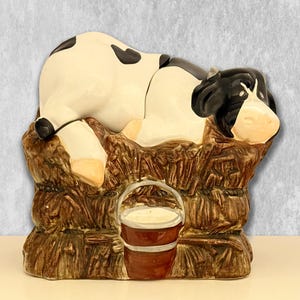 May include: A ceramic figurine of a black and white cow lying down on a brown textured hay bale. A small brown bucket with a silver rim is attached to the front of the hay bale. The cow has a white face with black patches around the eyes.