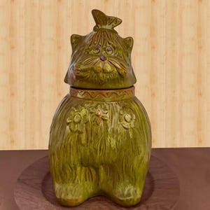 May include: A whimsical, green ceramic cat-shaped cookie jar with a lid featuring a butterfly. The jar is adorned with floral details and has a textured, fur-like appearance. The cat's face has purple eyes and a small, round nose.