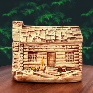 May include: A ceramic cabin-shaped container with a textured roof and log-cabin style walls. The container has a small door, two windows, and a chimney. The color palette includes shades of brown, beige, and cream.