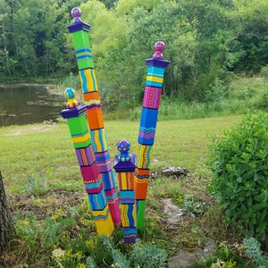May include: A cluster of colorful, painted wooden posts with decorative finials. The posts are painted in vibrant hues of green, yellow, orange, pink, and blue, with geometric and abstract patterns. They stand in a garden setting with a pond in the background.