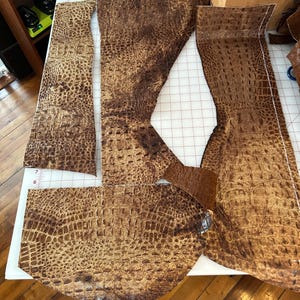 May include: Several pieces of brown crocodile-textured leather, cut into various shapes, are laid out on a white cutting mat. The leather displays a detailed, scale-like pattern and different shades of brown.