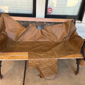 May include: A large, irregular piece of smooth, medium-brown leather draped over a park bench. The hide features a natural, matte finish with visible folds and creases across its surface, showcasing the organic shape and texture of the material.