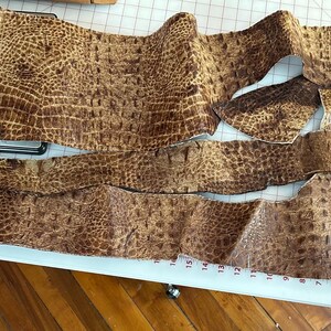 May include: Several pieces of brown faux crocodile leather, cut into different shapes, are laid out on a white cutting mat. The leather has a textured finish, possibly for crafting or design purposes.