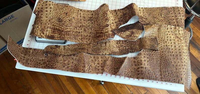May include: Several pieces of brown faux crocodile leather, cut into different shapes, are laid out on a white cutting mat. The leather has a textured finish, possibly for crafting or design purposes.