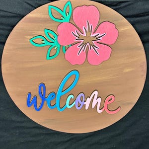 Może przedstawiać: Okrągły drewniany znak powitalny z różowym kwiatem i zielonymi liśćmi. Słowo "welcome" jest napisane kolorowym pismem. Znak ma brązowe malowane tło.