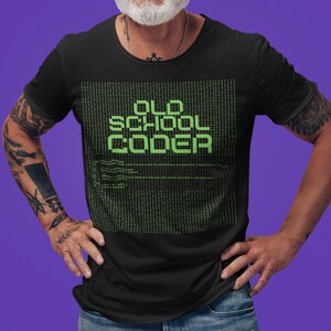 Può includere: T-shirt nero con la scritta "OLD SCHOOL CODER" in verde pixelato. Il design include uno sfondo a matrice e linee di codice. La maglietta è indossata da una persona tatuata.