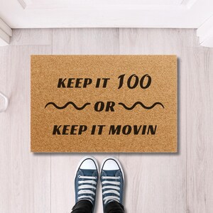 May include: A rectangular doormat made of natural fibers with the text "KEEP IT 100 OR KEEP IT MOVIN" in black. The doormat is placed in front of a pair of blue canvas sneakers.