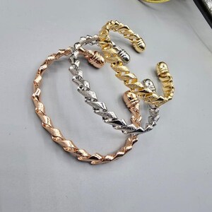 Può includere: Tre braccialetti a polsino aperti in oro rosa, argento e oro. Ogni braccialetto presenta un design a corda intrecciata. I braccialetti sono esposti su una superficie grigio chiaro, mettendo in risalto la loro lucentezza metallica e la trama complessa.