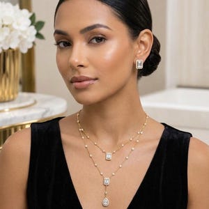 May include: A gold-toned layered necklace featuring small round clear stones along the chains, two rectangular pendants, and a single pear-shaped drop pendant. Matching rectangular stud earrings with clear stones are worn on the earlobes.