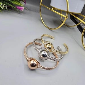 Può includere: Tre braccialetti a polsino aperti in oro, argento e oro rosa. Ogni braccialetto presenta una fascia strutturata e una grande sfera liscia al centro. I braccialetti sono esposti su una superficie grigio chiaro.