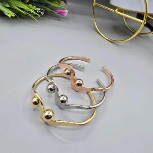 Può includere: Tre braccialetti a polsino aperti in oro, argento e oro rosa. Ogni braccialetto presenta un design curvo con un piccolo accento rotondo. I braccialetti sono esposti su una superficie grigio chiaro, con verde e un elemento decorativo dorato sullo sfondo.