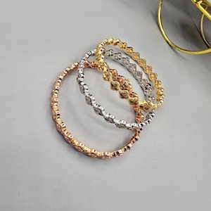 Può includere: Tre delicati braccialetti in oro rosa, argento e oro. Ogni braccialetto presenta un motivo floreale ripetuto. I braccialetti sono esposti su una superficie grigio chiaro, con un oggetto dorato sullo sfondo.