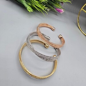 Può includere: Tre braccialetti a polsino in metallo dorato, argentato e oro rosa. I braccialetti hanno un design intrecciato e estremità arrotondate. Il braccialetto dorato è in basso, quello argentato al centro e quello oro rosa in alto. I braccialetti sono esposti su una superficie grigia.