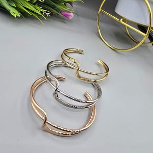 Può includere: Sono presentati tre braccialetti a polsino aperti in oro, argento e oro rosa. I braccialetti hanno un design minimalista con una forma curva e aperta. Un oggetto decorativo dorato è sullo sfondo, insieme a del verde e fiori viola.