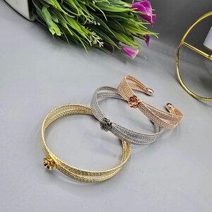 Può includere: Tre braccialetti in metallo dorato, argentato e oro rosa. Ogni braccialetto presenta un motivo floreale. I braccialetti sono esposti su una superficie grigia, con verde e fiori viola sullo sfondo.