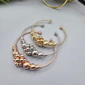 Può includere: Tre braccialetti a polsino aperti in oro, argento e oro rosa. Ogni braccialetto presenta un design a perline con trame e dimensioni variabili. I braccialetti sono esposti su una superficie grigia, mettendo in mostra le loro diverse finiture metalliche.