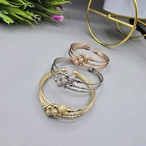 Può includere: Tre braccialetti a polsino aperti in oro, argento e oro rosa. Ogni braccialetto presenta un gruppo di abbellimenti rotondi e scintillanti. I braccialetti sono esposti su una superficie grigio chiaro, mostrando il loro design elegante.