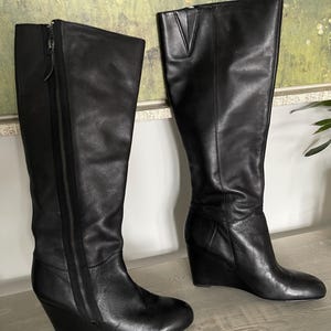 May include: A pair of black leather knee-high wedge boots. One boot features a full-length zipper on the side. The boots have a wedge heel and a rounded toe. The boots are displayed on a light gray surface.