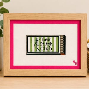 May include: A framed needlepoint featuring the text "Lex Stitch Club" in green and white, set against a striped background. The artwork is framed in a light wood frame with a hot pink mat.