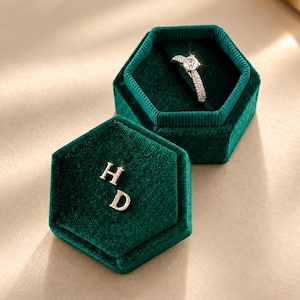 May include: A hexagonal dark green velvet ring box with a removable lid. The lid features the letters H and D in a metallic finish. Inside the box, a silver-toned ring with a central round-cut stone and a pavé-set band is displayed upright.