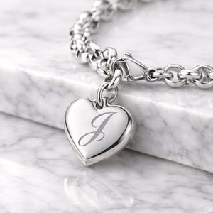 Könnte beinhalten: Ein silbernes Armband mit einem herzförmigen Anhänger, auf dem der Buchstabe "J" eingraviert ist. Das Armband besteht aus verbundenen, abgerundeten Silberstücken. Der Hintergrund ist eine weiße Marmoroberfläche.