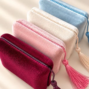 May include: Four rectangular velvet pouches arranged in a row, featuring colors of deep burgundy, soft pink, cream, and light blue. Each pouch is finished with a matching twisted cord trim along the top edge and a decorative hanging tassel.