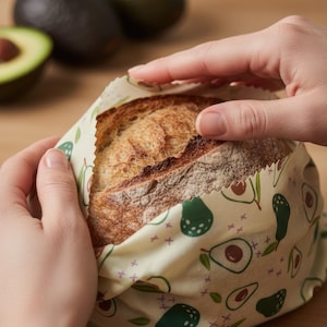 May include: A loaf of bread wrapped in a reusable beeswax food wrap. The wrap features a cream background with a pattern of green and brown avocados and purple crosses. The bread is held by two hands.