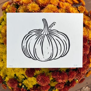 May include: A rectangular white paper print featuring a black ink linocut illustration of a pumpkin with a stem and a curly vine. The bottom left corner displays the text 2026, and the bottom right corner features the initials BER in black ink.