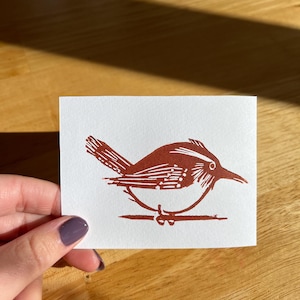 Puede incluir: Una pequeña tarjeta rectangular con una impresión en linóleo marrón rojizo de un pájaro posado en una rama. El pájaro tiene marcas detalladas en las alas y el cuerpo. El fondo es blanco y la tarjeta se sostiene sobre una superficie de madera.
