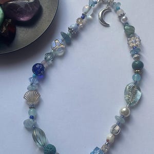 May include: A phone charm featuring a variety of blue, clear, and white beads, including spherical, faceted, and textured shapes. The charm includes a silver-toned crescent moon pendant and a black cord loop for attachment to a mobile device.