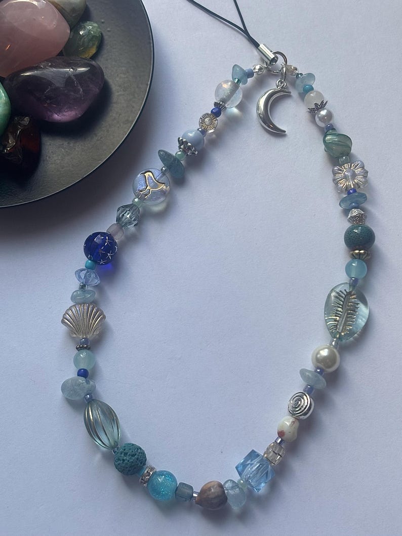 May include: A phone charm featuring a variety of blue, clear, and white beads, including spherical, faceted, and textured shapes. The charm includes a silver-toned crescent moon pendant and a black cord loop for attachment to a mobile device.