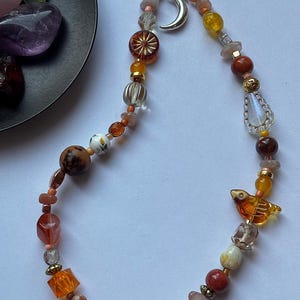 May include: A beaded phone charm featuring a variety of orange, brown, and clear beads in different shapes and textures. The charm includes a silver crescent moon pendant and a small amber-colored bird-shaped bead, all strung on a black cord.