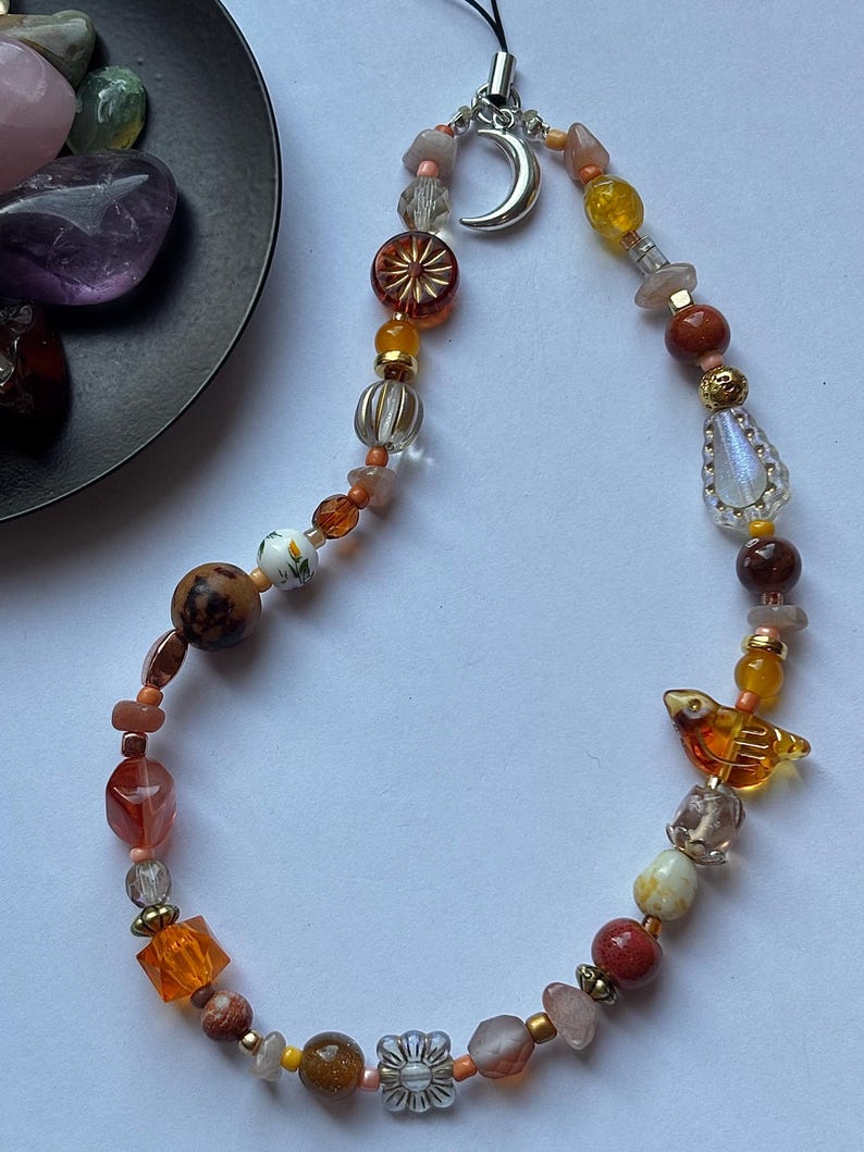 May include: A beaded phone charm featuring a variety of orange, brown, and clear beads in different shapes and textures. The charm includes a silver crescent moon pendant and a small amber-colored bird-shaped bead, all strung on a black cord.