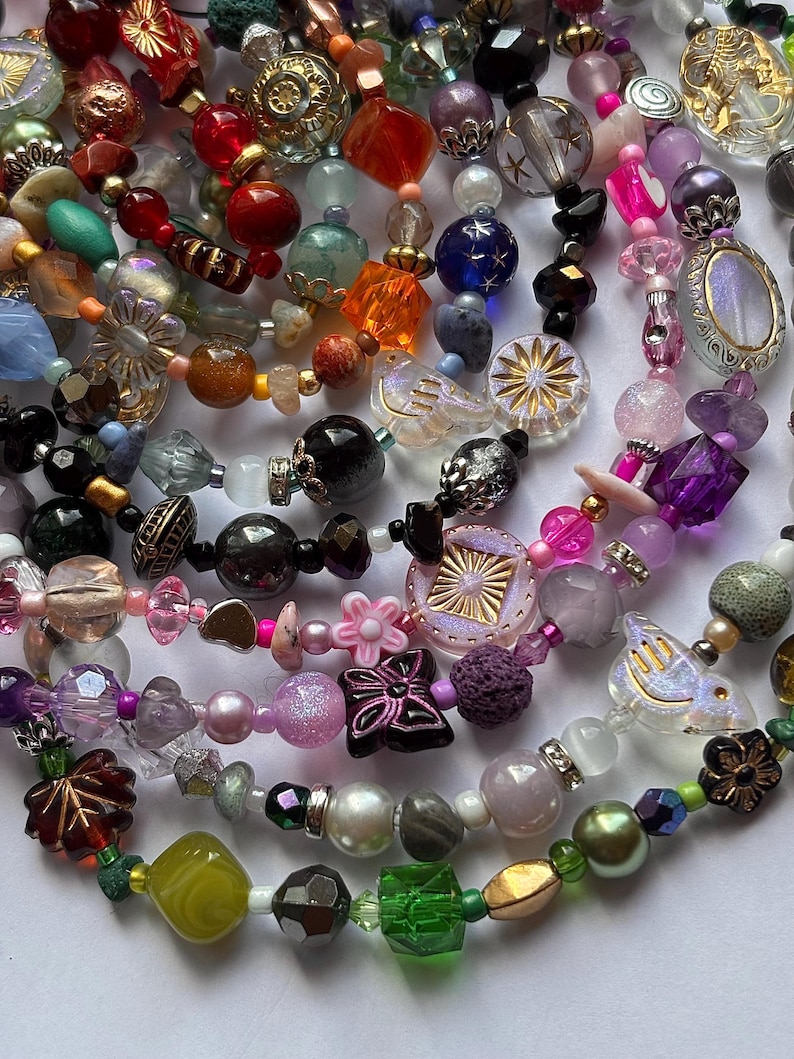 May include: A collection of colorful beaded necklaces with various shapes and sizes. The beads are in shades of red, orange, blue, purple, pink, green, and gold. Some beads have floral and starburst designs, and others are iridescent.