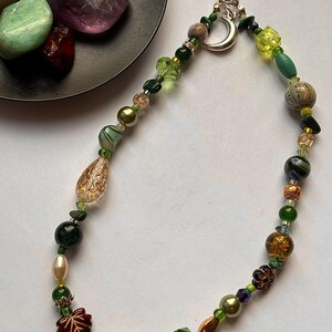 May include: A phone charm strap featuring a variety of beads in shades of green, brown, and gold. The design includes a silver crescent moon charm, a clear teardrop bead, a dark red leaf-shaped bead, and assorted glass, pearl, and stone beads.