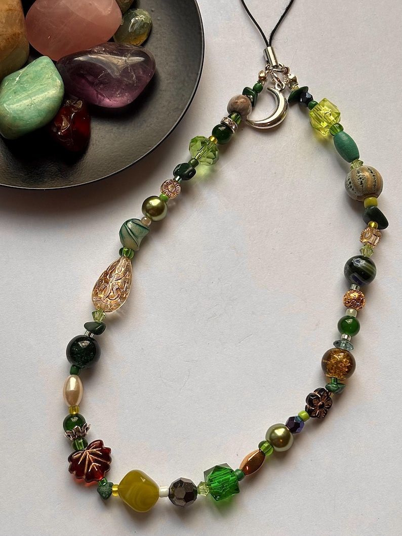 May include: A phone charm strap featuring a variety of beads in shades of green, brown, and gold. The design includes a silver crescent moon charm, a clear teardrop bead, a dark red leaf-shaped bead, and assorted glass, pearl, and stone beads.