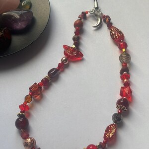 May include: A phone charm featuring a variety of red beads in different shapes, sizes, and textures, including faceted, smooth, and patterned glass beads. A silver-toned crescent moon charm is attached to the top, connected to a black cord loop.